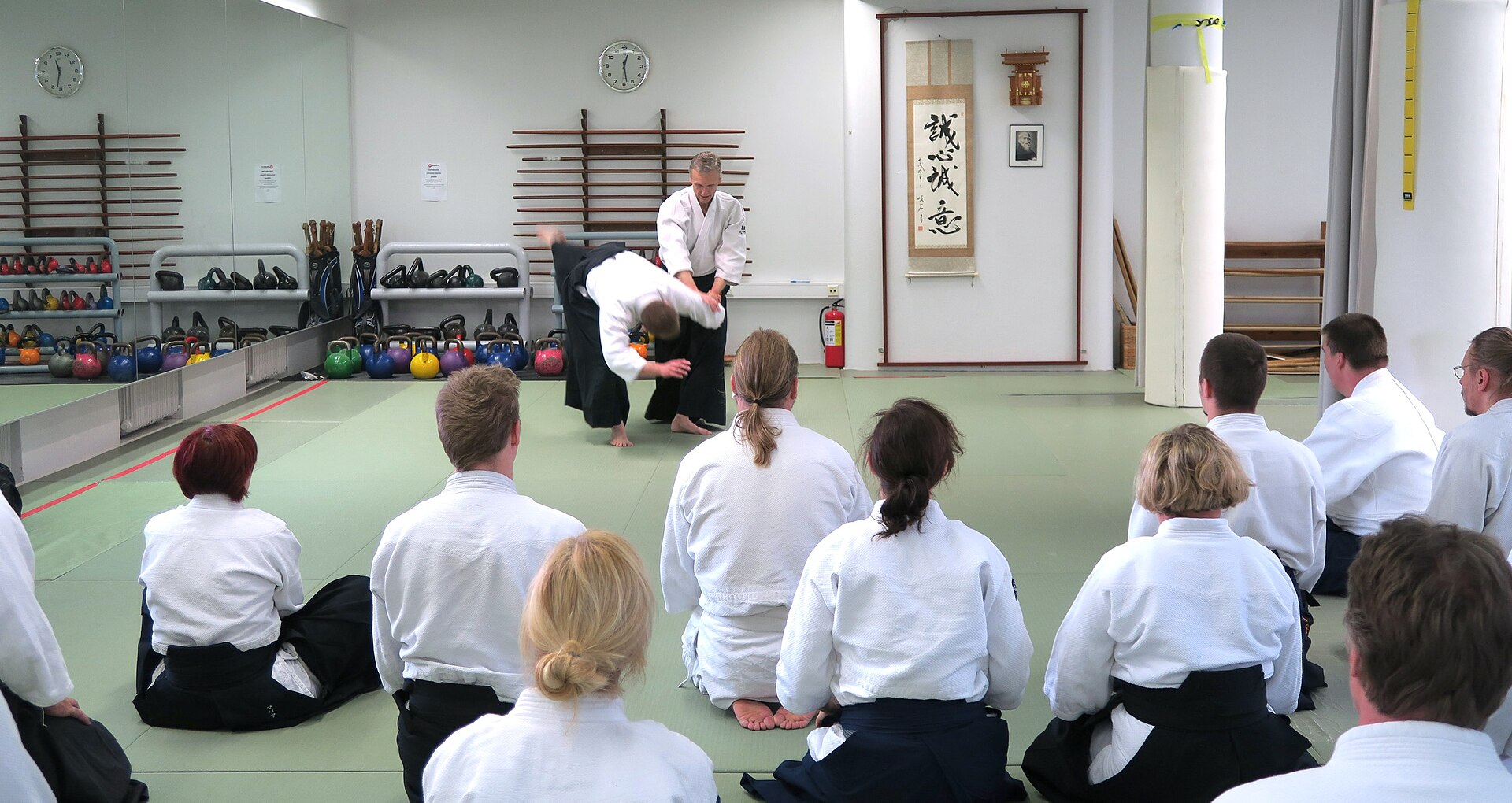 Un enseignant présente un mouvement d’aïkido devant un groupe de pratiquants adultes assis dans un dojo